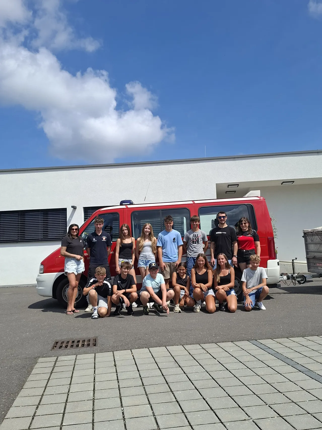 Eine Gruppe von Teenagern und jungen Erwachsenen posiert für ein Foto vor einem roten Van. Sie stehen und knien auf einem gepflasterten Bereich. Dahinter steht ein weißes Gebäude mit einem bewölkten Himmel.