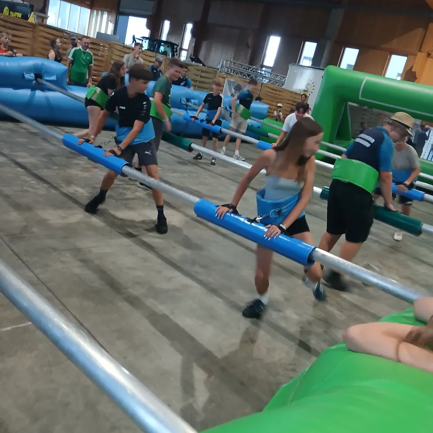 Eine Gruppe junger Menschen nimmt an einem Indoor-Hindernisparcours teil, hält sich an blauen Stangen fest und navigiert durch aufblasbare Hindernisse.