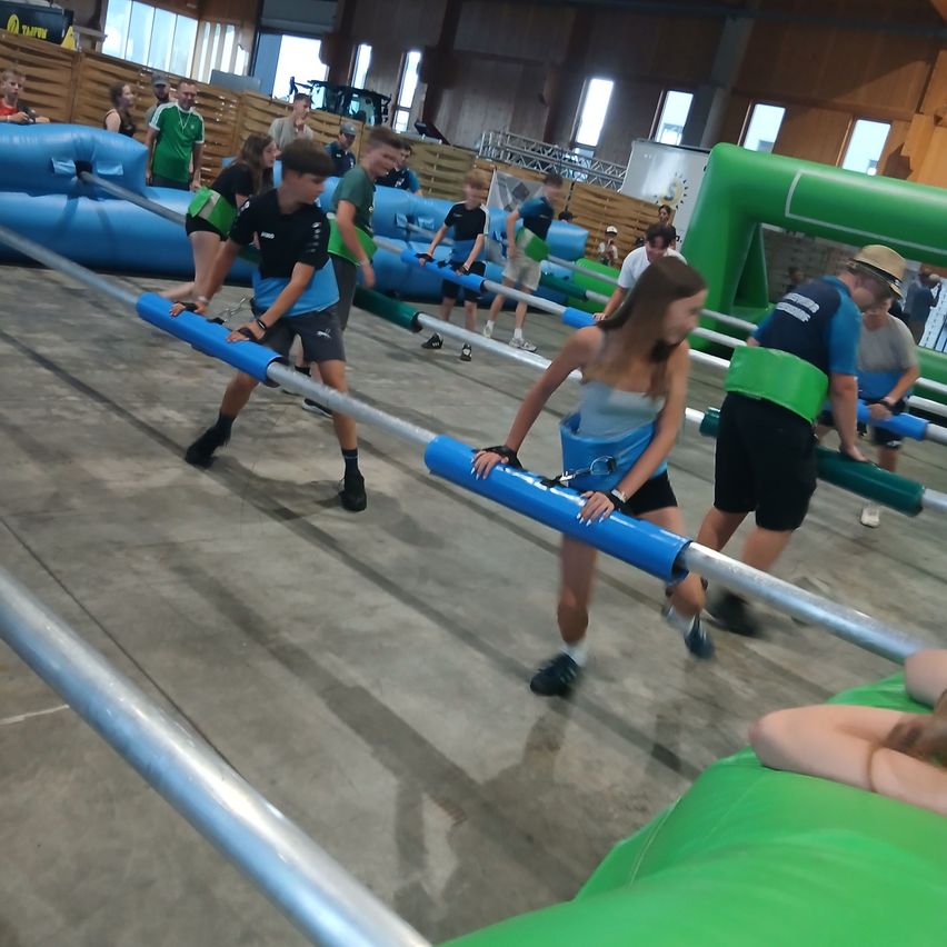 Eine Gruppe junger Menschen nimmt an einem Indoor-Hindernisparcours teil, hält sich an blauen Stangen fest und navigiert durch aufblasbare Hindernisse.