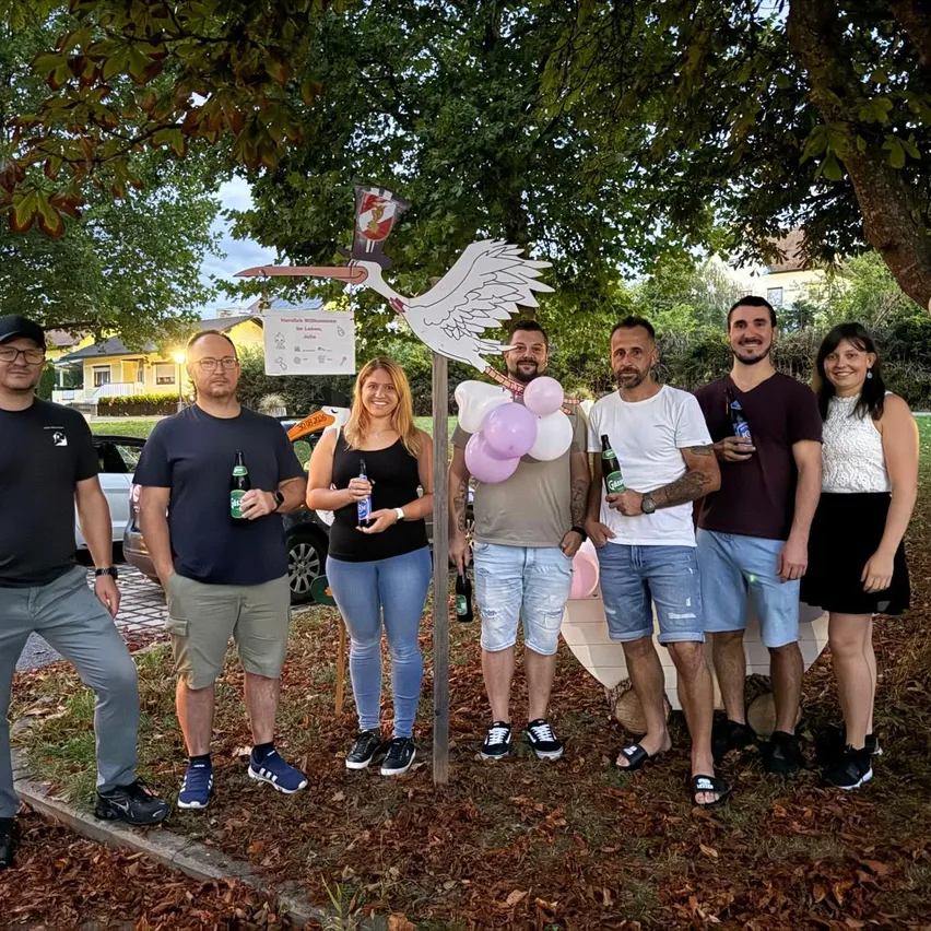 Eine Gruppe von Menschen steht unter einem Baum mit einem Storch-Ausschnitt und Luftballons und hält Flaschen Bier. Sie sind lässig gekleidet und scheinen für ein Foto zu posieren.