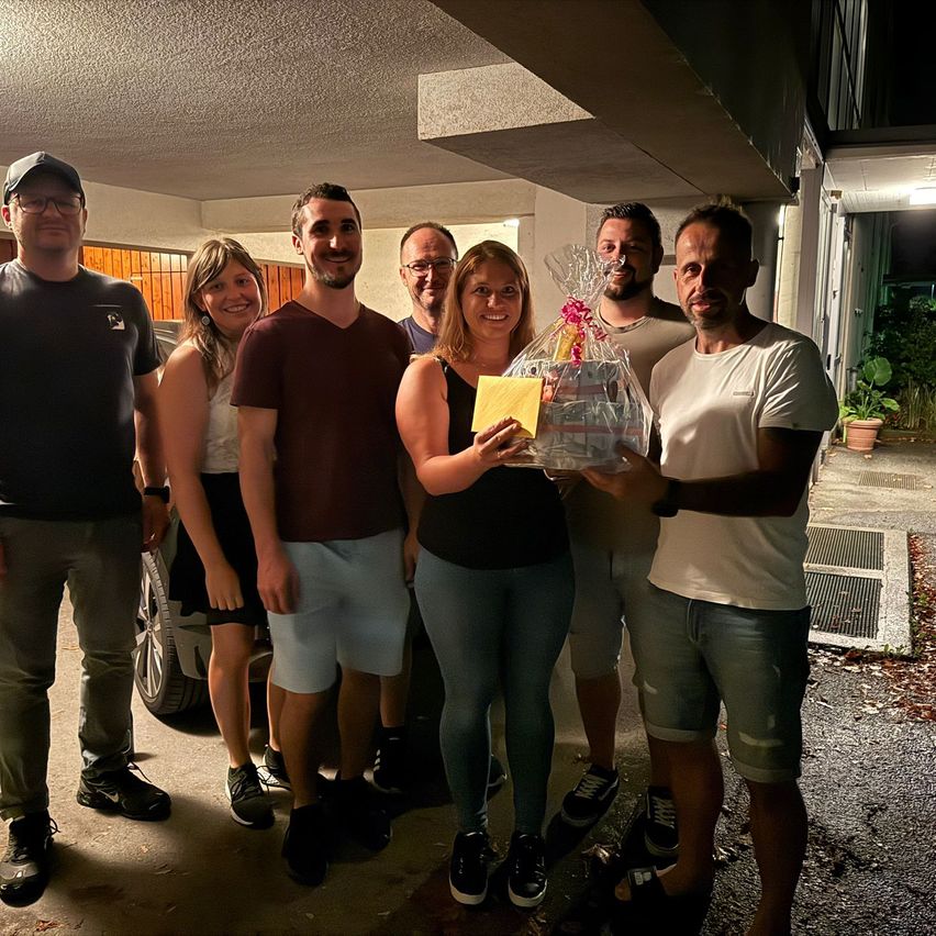 Eine Gruppe von Menschen steht in einer Garage, lächelt und hält ein verpacktes Geschenk. Die Frau in der Mitte hält eine gelbe Karte.