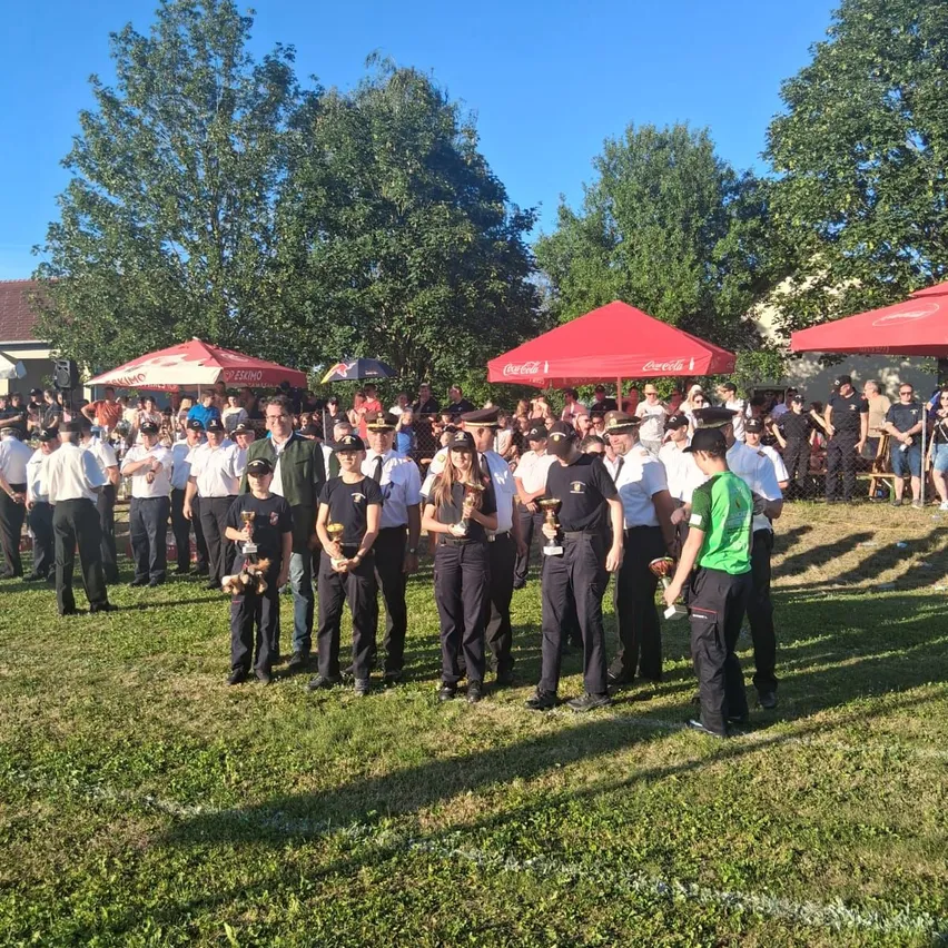 Eine Gruppe von Menschen steht auf einem Feld und hält Pokale. Sie tragen Uniformen und Hüte. Dahinter versammelt sich eine Menschenmenge unter Schirmen. Bäume und ein Haus sind im Hintergrund.