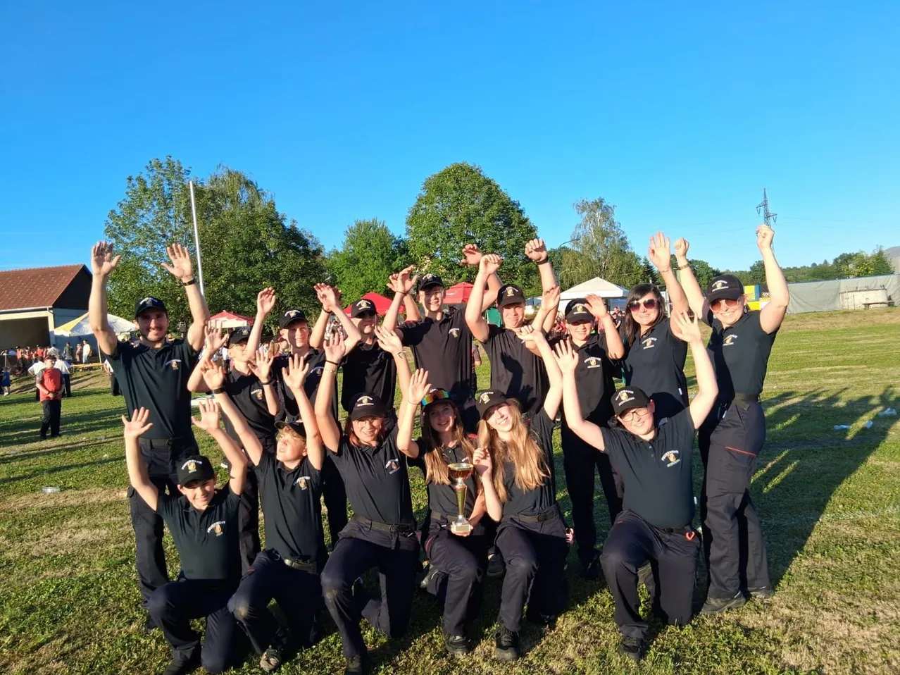 Ein Team in schwarzen Uniformen und Mützen posiert für ein Foto, hebt die Hände und hält einen Pokal. Sie stehen auf Gras mit Bäumen und Zelten im Hintergrund.