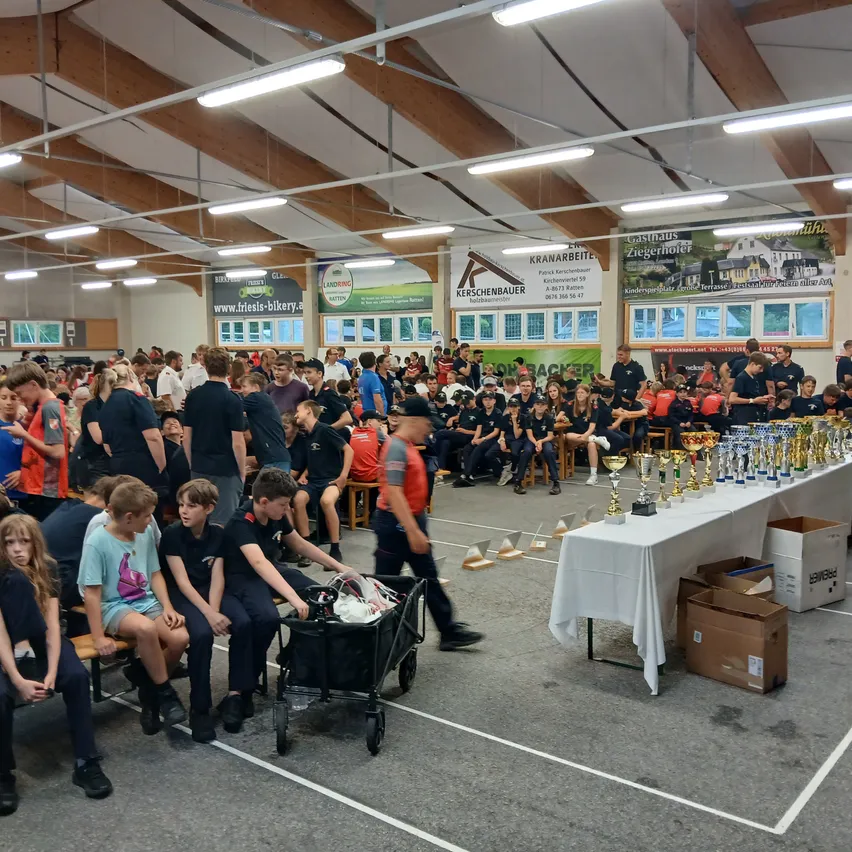 Eine Gruppe von Menschen ist in einem Saal versammelt und schaut sich einen Wettbewerb an. Einige sitzen auf Bänken, andere stehen herum. Es gibt Trophäen auf einem Tisch zur Schau.