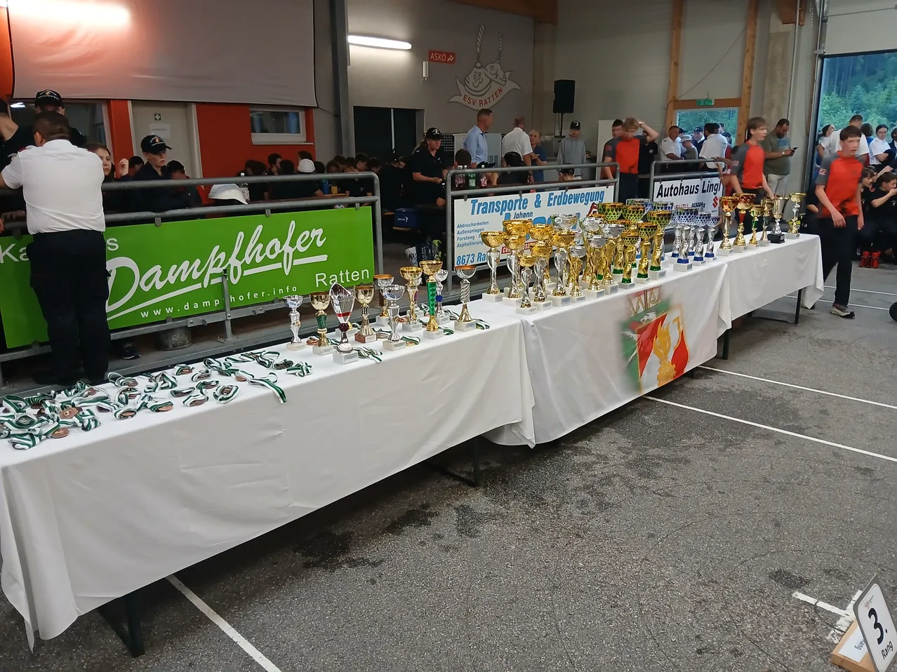 Eine Outdoor-Veranstaltung mit Menschen hinter einem Zaun. Davor steht ein mit weißem Tuch bedeckter Tisch mit Trophäen und Medaillen.