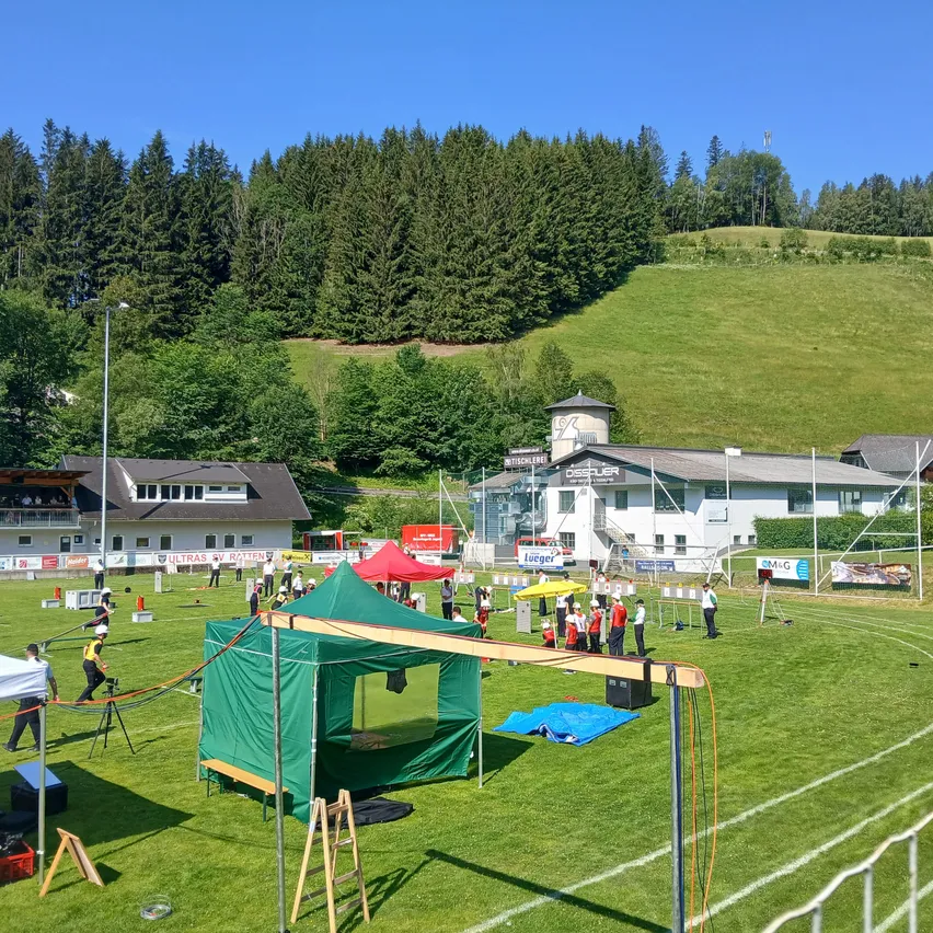 Ein grünes Zelt und mehrere Personen befinden sich auf einem Rasenfeld. Es gibt Banner und Zelte in der Umgebung. In der Ferne gibt es Gebäude und einen mit Bäumen bedeckten Hügel.