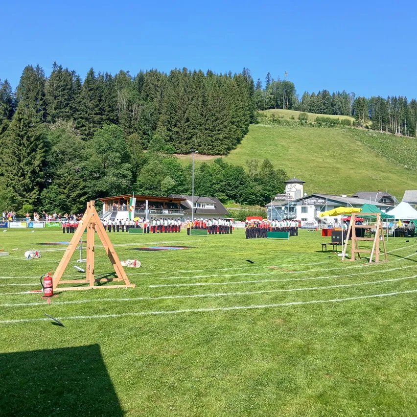 Ein Sportplatz mit einem Feuerlöscher und einem dreieckigen Ziel. Mehrere Personen stehen in Reihen. Einige Zelte sind auf dem Feld aufgestellt. In der Ferne befinden sich viele Bäume und ein Gebäude.