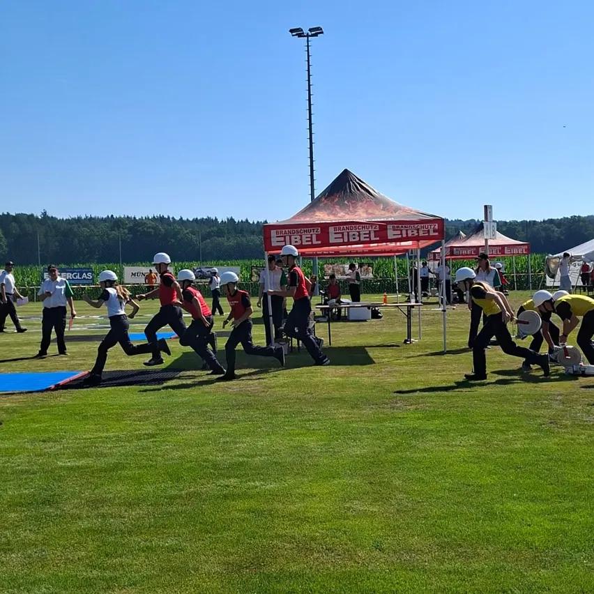 Eine Gruppe von Menschen in übereinstimmenden roten Uniformen und Helmen nimmt an einer Laufaktivität auf einem Feld teil, mit Zelten und einem Tisch im Hintergrund.