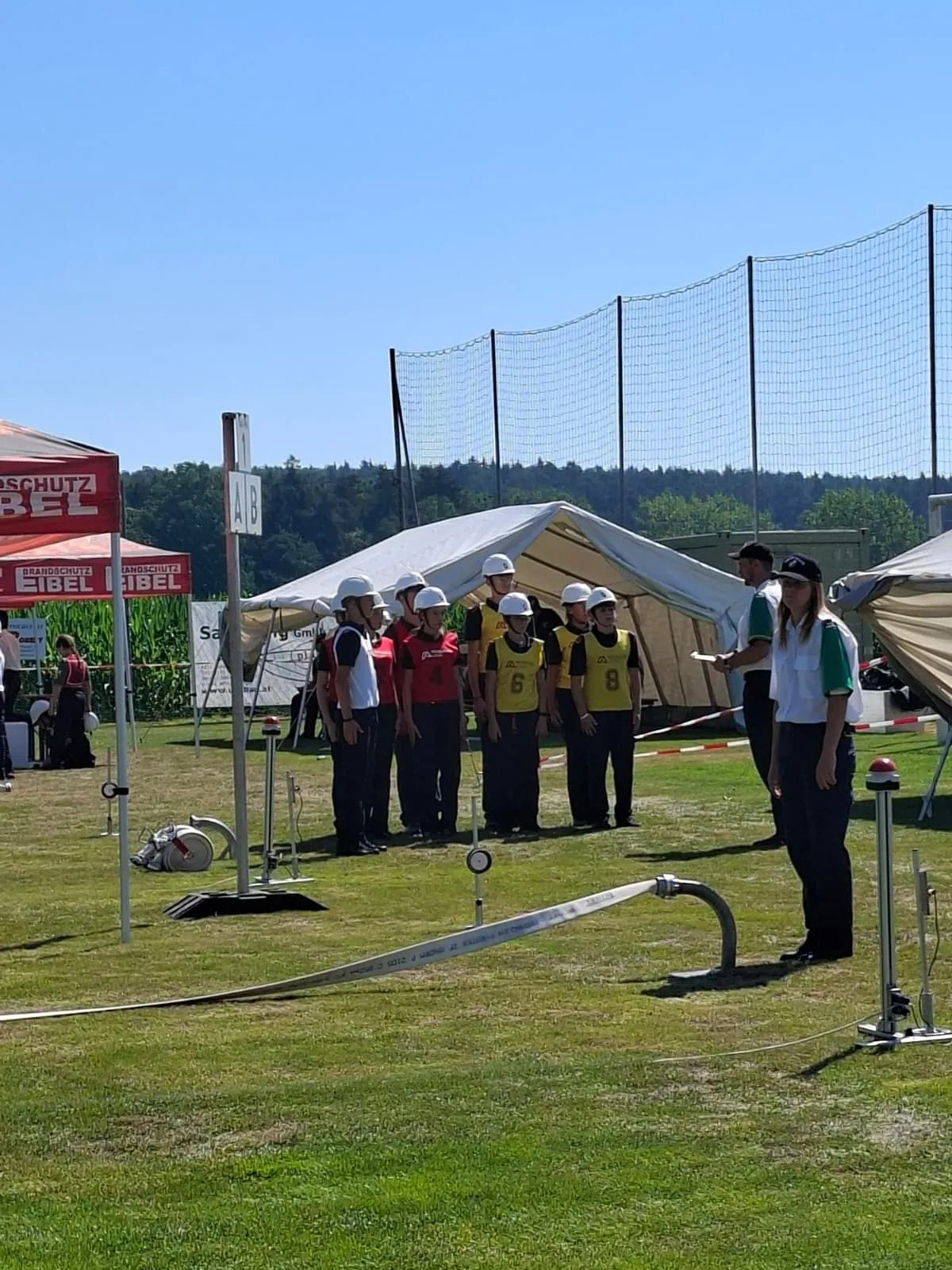 Eine Gruppe von Menschen in nummerierten Trikots und Helmen steht auf einem Feld, wahrscheinlich bei einer Sportveranstaltung. Zelte und ein Netzzaun sind im Hintergrund zu sehen.