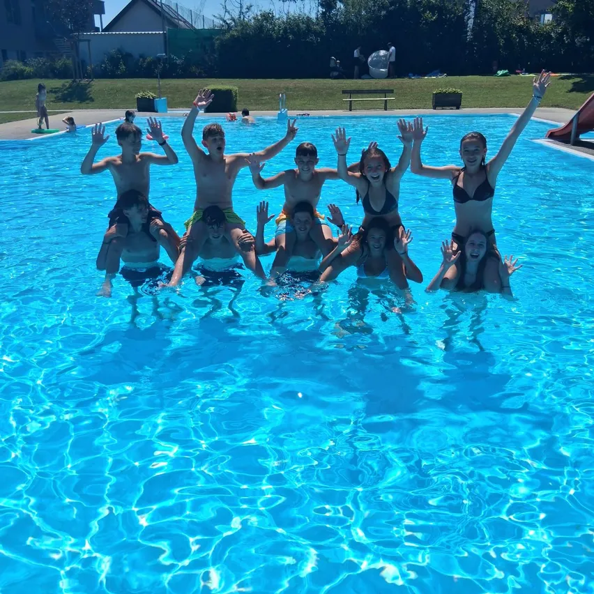 Eine Gruppe von Teenagern posiert im Schwimmbecken mit erhobenen Händen. Einige tragen Badeanzüge, andere T-Shirts. Eine Rutsche ist im Hintergrund zu sehen.