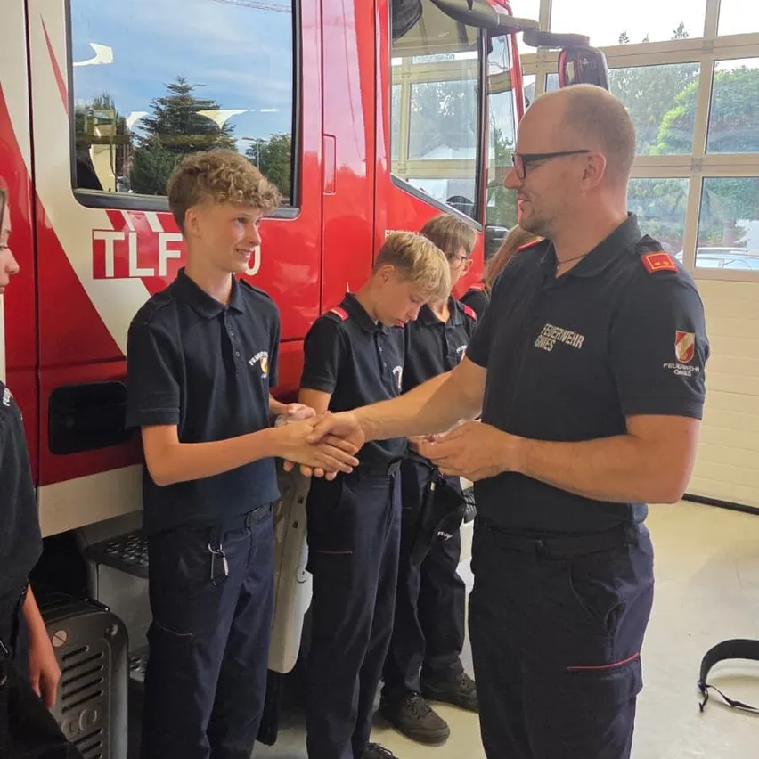 Zwei junge Jungen in dunkelblauen Uniformen schütteln die Hände eines Feuerwehrmanns vor einem roten Feuerwehrauto.
