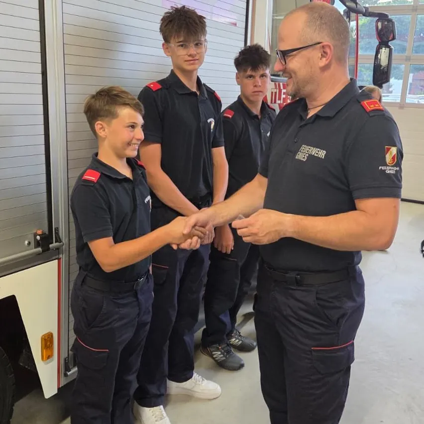 Ein Feuerwehrmann gibt einem jungen Jungen in Uniform die Hand, wobei zwei andere Jungen hinter ihnen in einer Feuerwache stehen.