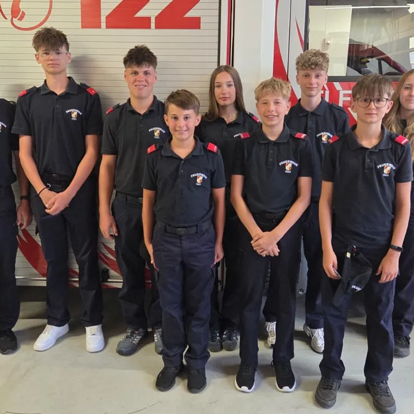 Eine Gruppe junger Personen in schwarzen Uniformen, möglicherweise Feuerwehrleute, steht vor einem Feuerwehrwagen.