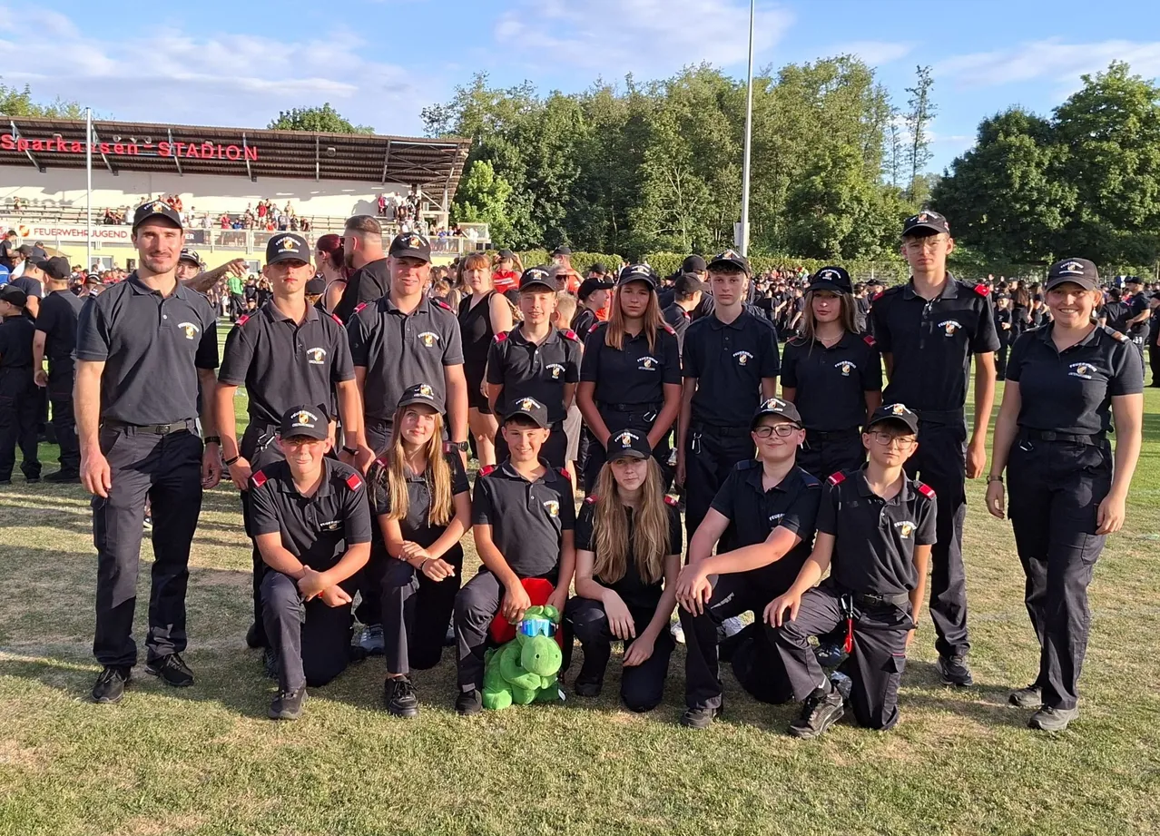 Eine Gruppe von Menschen in Uniform posiert für ein Foto auf einem Feld. Die Gruppe besteht aus jungen Männern und Frauen mit Mützen und Turnschuhen. Eine grüne Puppe ist in der Mitte platziert.