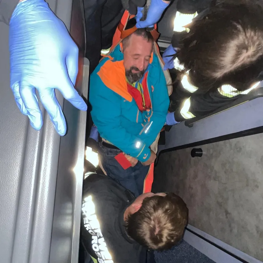 Ein Mann in einer blauen Jacke und Jeans wird von zwei Personen in einem Fahrzeug unterstützt.
