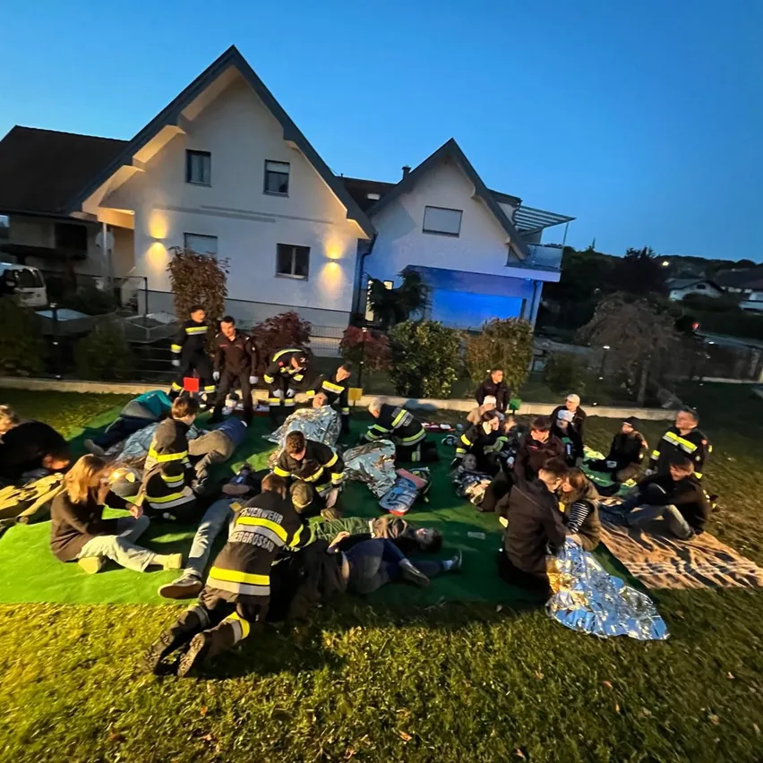 Eine Gruppe von Feuerwehrleuten sitzt auf dem Rasen vor einem Haus, einige von ihnen tragen Helme und Jacken.