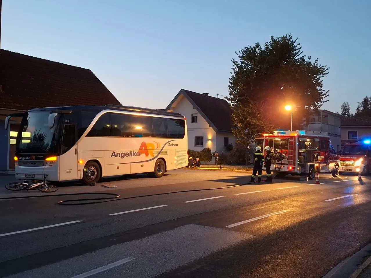 Ein weißer Bus mit dem Wort Angelika an der Seite ist am Straßenrand geparkt. Ein Feuerwehrwagen ist daneben geparkt, mit zwei Feuerwehrleuten, die daneben stehen.