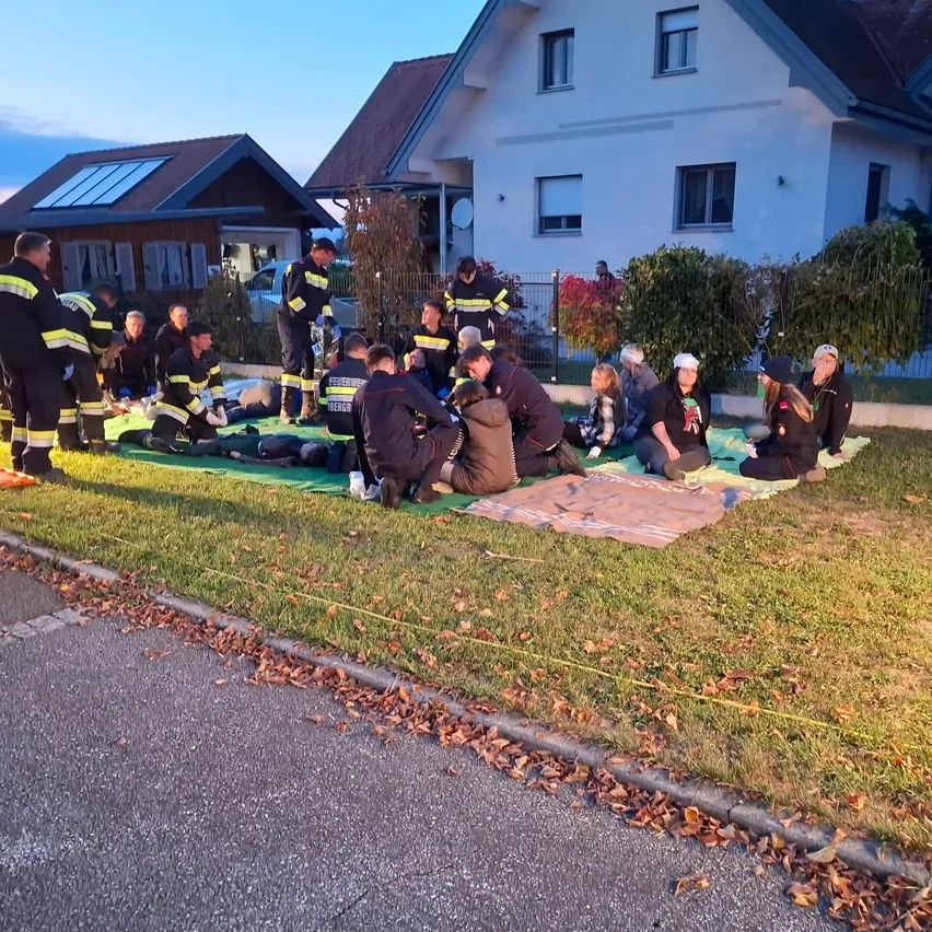 Eine Gruppe von Feuerwehrleuten und Menschen sitzt auf dem Rasen vor einem Haus. Einige von ihnen tragen Hüte und Jacken.