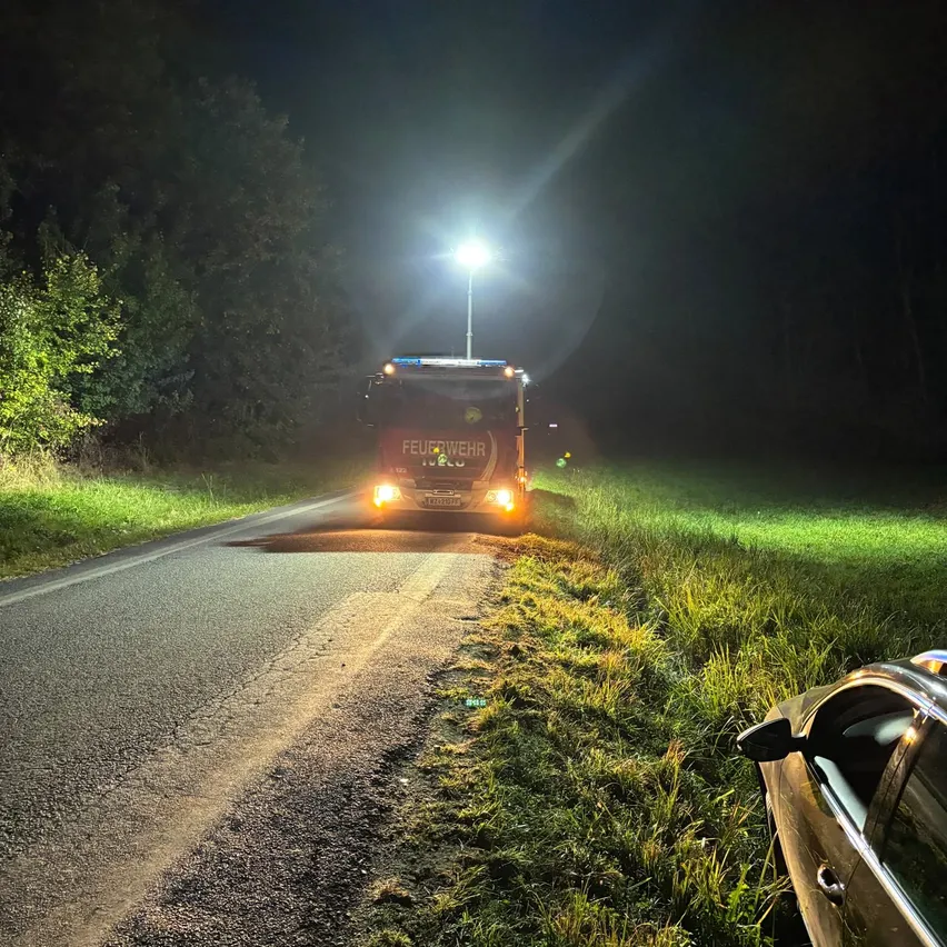 Ein Feuerwehrauto fährt nachts auf einer Straße. Die Scheinwerfer sind eingeschaltet und beleuchten den Weg vor dem Fahrzeug.