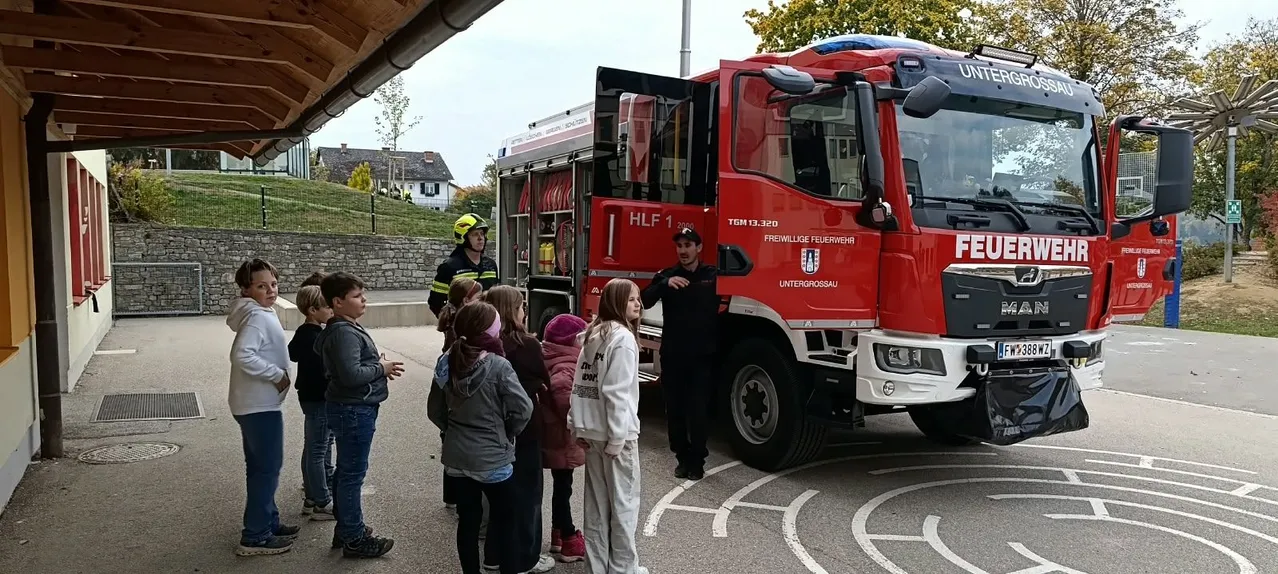 Eine Gruppe von Kindern steht vor einem roten Feuerwehrauto mit zwei Feuerwehrleuten, von denen einer einen gelben Helm trägt.