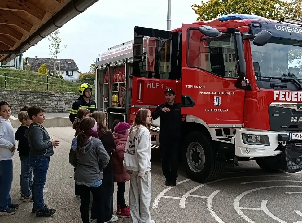 Eine Gruppe von Kindern steht vor einem roten Feuerwehrauto mit zwei Feuerwehrleuten, von denen einer einen gelben Helm trägt.