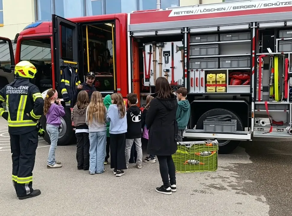 Eine Gruppe von Kindern steht um einen Feuerwehrwagen mit offenen Türen herum. Ein Feuerwehrmann steht vor ihnen und erklärt etwas.