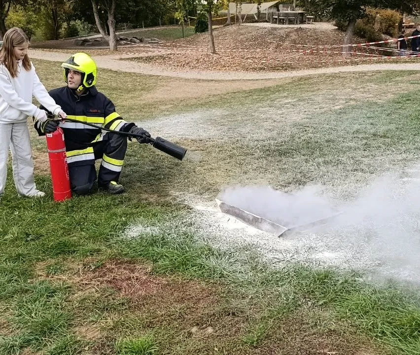 Ein Feuerwehrmann in voller Ausrüstung verwendet einen Feuerlöscher, um ein kleines Feuer auf einem Grasfeld zu löschen. Ein Mädchen in Weiß steht in der Nähe und beobachtet.