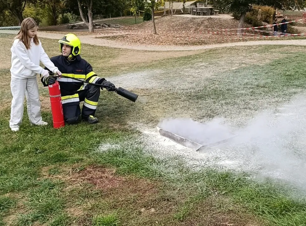 Ein Feuerwehrmann in voller Ausrüstung verwendet einen Feuerlöscher, um ein kleines Feuer auf einem Grasfeld zu löschen. Ein Mädchen in Weiß steht in der Nähe und beobachtet.