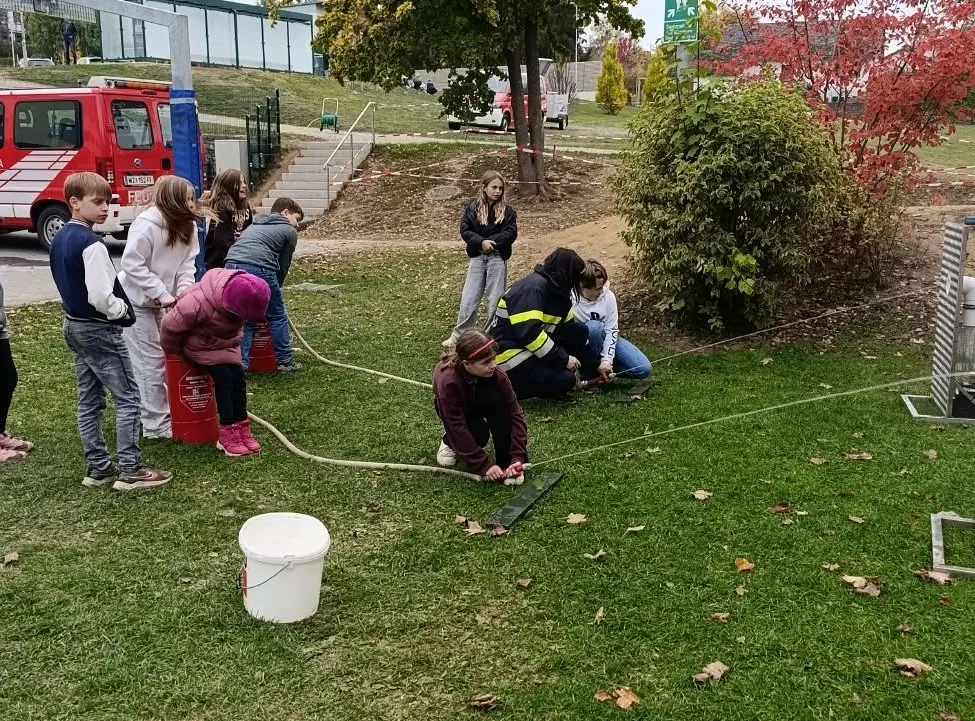 Eine Gruppe von Kindern und Erwachsenen versammelt sich auf einer Rasenfläche, wobei einige von ihnen einen Schlauch halten und andere herumstehen. Ein Feuerwehrauto ist in der Nähe geparkt, und es gibt Bäume und Pflanzen im Hintergrund.