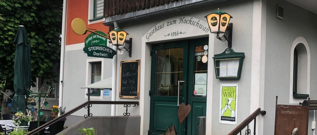 Ein traditionelles deutsches Gasthaus mit einem hölzernen Balkon, einer grünen Tür und blumengeschmückten Stufen. Das Schild lautet Gasthaus zum Hochschwab, und es gibt verschiedene Pflanzen und Dekorationen rund um den Eingang.
