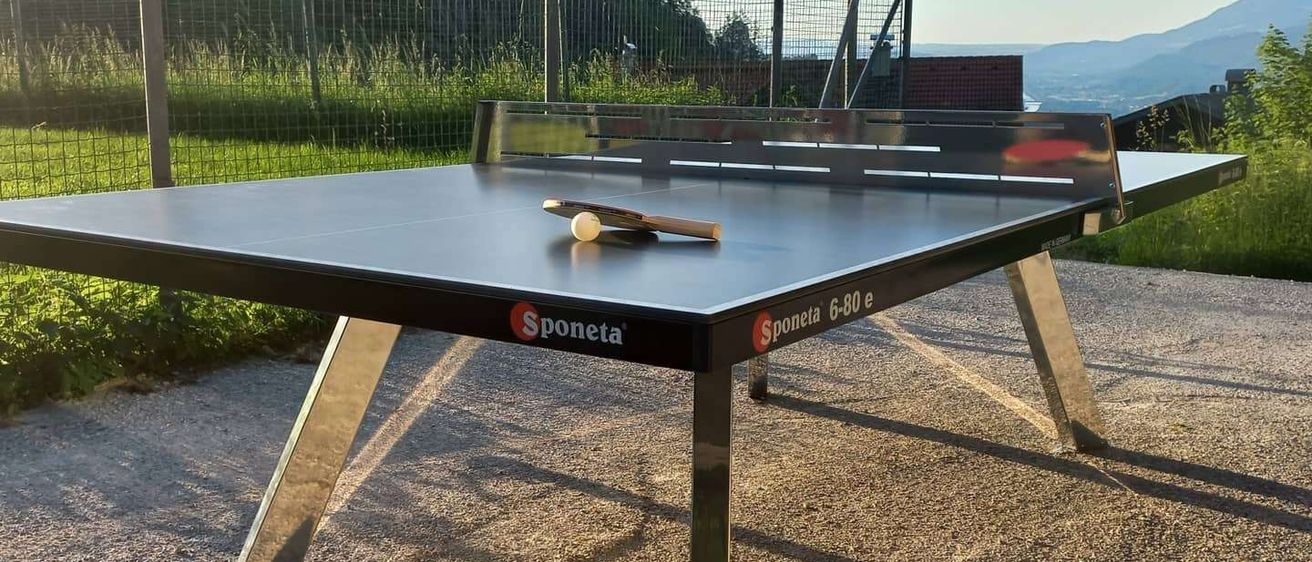 Ein schwarzer Outdoor-Tischtennistisch mit Sponeta-Marke steht auf Kies. Ein Tischtennisschläger und ein Ball liegen auf dem Tisch. Dahinter befindet sich ein Zaun mit einer Rasenfläche und Bergen.