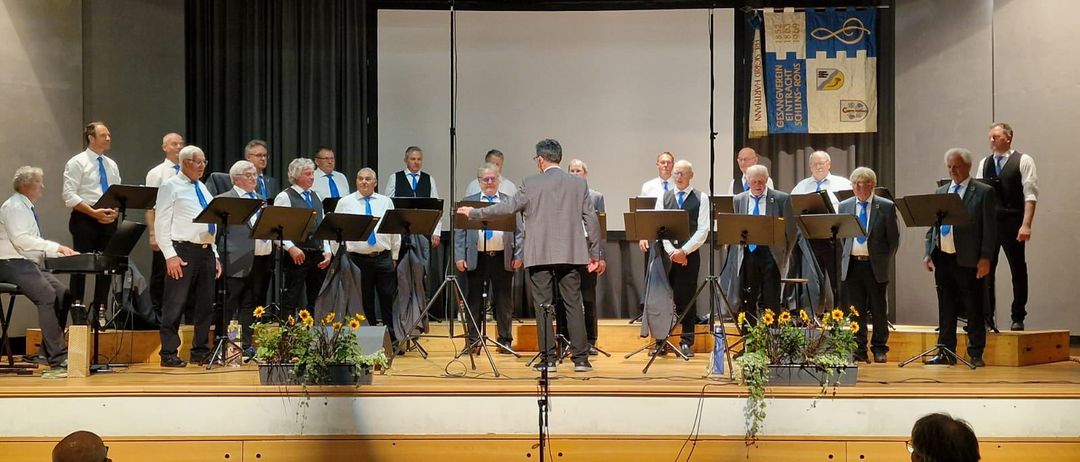 Ein Chor tritt auf der Bühne mit einem Podium auf, gekleidet in formelle Kleidung. Die Bühne ist mit Musikständern und Blumen geschmückt. Im Hintergrund sind ein Bildschirm und Banner zu sehen.