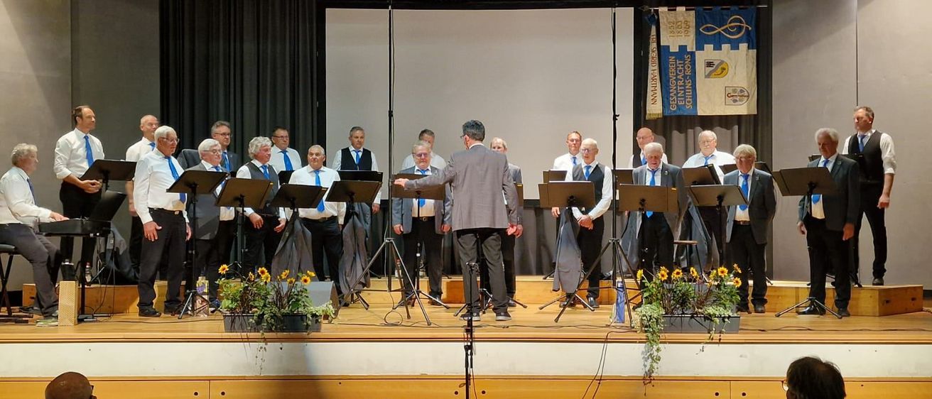 Ein Chor tritt auf der Bühne mit einem Podium auf, gekleidet in formelle Kleidung. Die Bühne ist mit Musikständern und Blumen geschmückt. Im Hintergrund sind ein Bildschirm und Banner zu sehen.