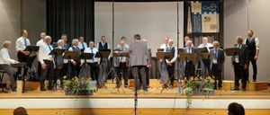 Ein Chor tritt auf der Bühne mit einem Podium auf, gekleidet in formelle Kleidung. Die Bühne ist mit Musikständern und Blumen geschmückt. Im Hintergrund sind ein Bildschirm und Banner zu sehen.