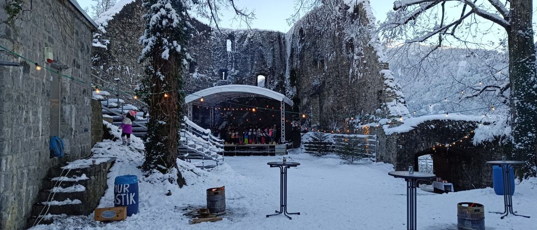 Verschneite Ruinen mit einer Bühneneinrichtung. Eine Gruppe von Menschen steht auf der Bühne. Vor der Bühne befinden sich Tische mit Flaschen. Ein Fass und ein Lagerbox sind in der Nähe.