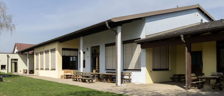 Ein modernes Gebäude mit einem braunen Dach und weißen Wänden, ausgestattet mit mehreren Fenstern und einer Terrasse mit Holzbänken und einem Tisch.