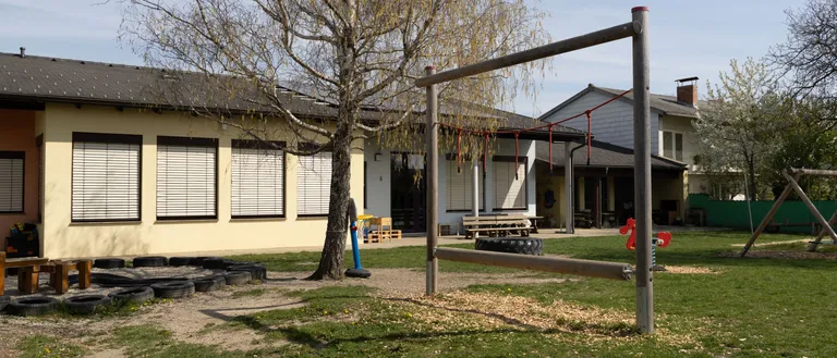 Ein Spielplatz mit einem Baum, einer Schaukel und einer Reifenstange vor einem Gebäude mit weißen Fensterläden.