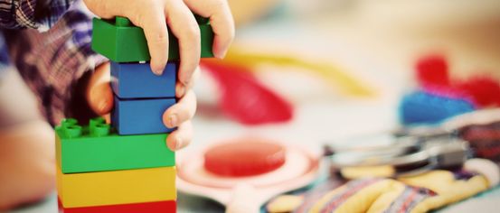 Ein Kind stapelt bunte Lego-Steine auf einem Tisch, mit verschiedenen Spielzeugen und einem Paar Handschuhen im Hintergrund.