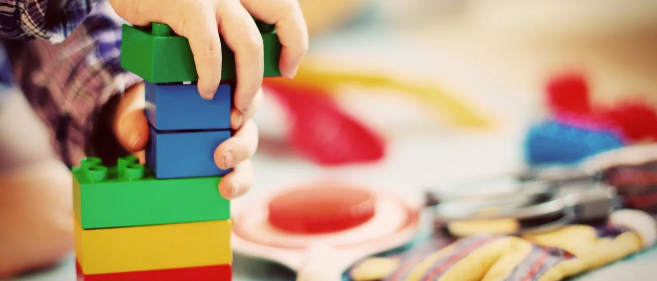 Ein Kind stapelt bunte Lego-Steine auf einem Tisch, mit verschiedenen Spielzeugen und einem Paar Handschuhen im Hintergrund.