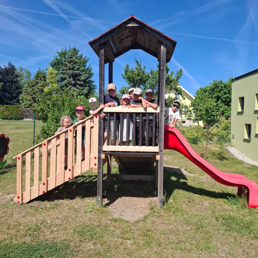 Eine Gruppe von Kindern spielt an einer hölzernen Spielstruktur mit einer roten Rutsche an einem sonnigen Tag.