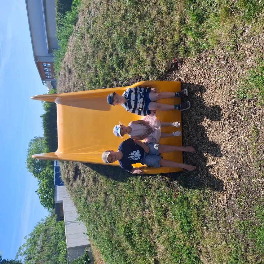 Drei Kinder sitzen auf einem gelben Rutschen auf einem Hügel. Eines von ihnen trägt einen Hut. Der Rutsch ist von grünem Gras und Bäumen umgeben.