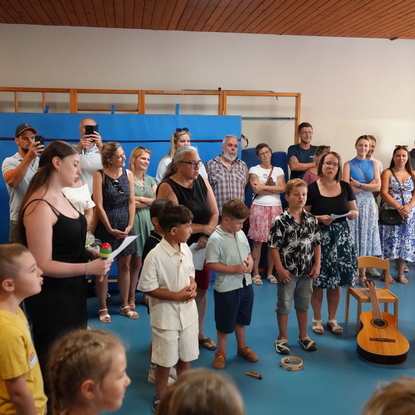 Eine Gruppe von Kindern und Erwachsenen versammelt sich in einem Raum. Einige Kinder halten Instrumente, und eine Frau hält ein Stück Papier. Der Raum hat eine blaue Wand und eine Holzdecke.