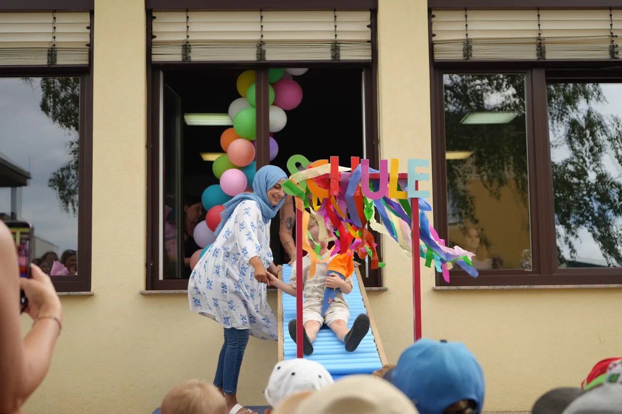 Eine Frau mit einem blauen Kopftuch hilft einem Kind, einen bunten Schlitten mit dem Wort 'Schule' darauf hinunterzurutschen. Hinter ihnen zeigt ein Fenster Luftballons und die Aussicht auf einen Baum.