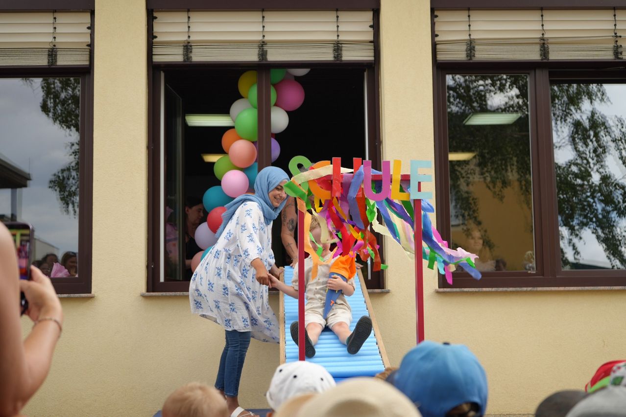 Eine Frau mit einem blauen Kopftuch hilft einem Kind, einen bunten Schlitten mit dem Wort 'Schule' darauf hinunterzurutschen. Hinter ihnen zeigt ein Fenster Luftballons und die Aussicht auf einen Baum.