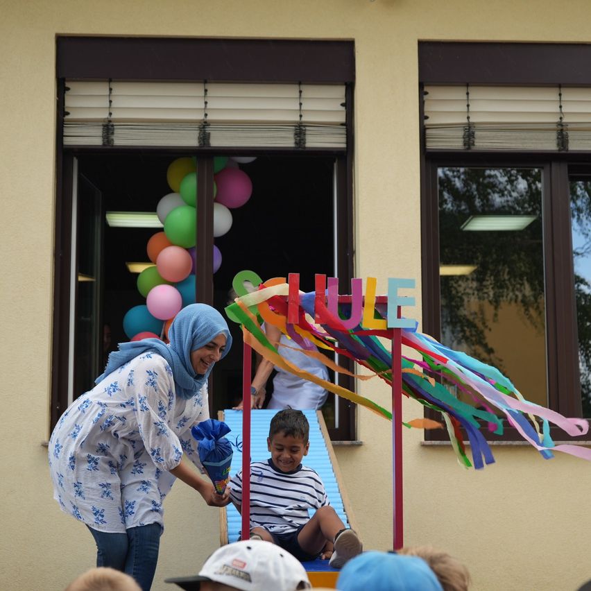 Eine Frau mit einem blauen Kopftuch überreicht einem Jungen, der in einem geschmückten Wagen sitzt, ein Geschenk vor einem Gebäude mit bunten Luftballons im Fenster.