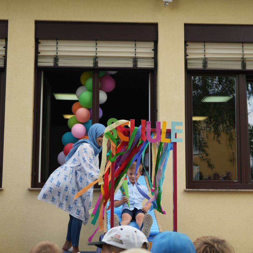 Eine Frau in einem blauen Kopftuch hilft einem kleinen Jungen in einem weißen Hemd und schwarzen Shorts, eine bunte Bandleiter vor einem Gebäude mit Luftballons und einem Schild mit der Aufschrift 'Schule' zu erklimmen.
