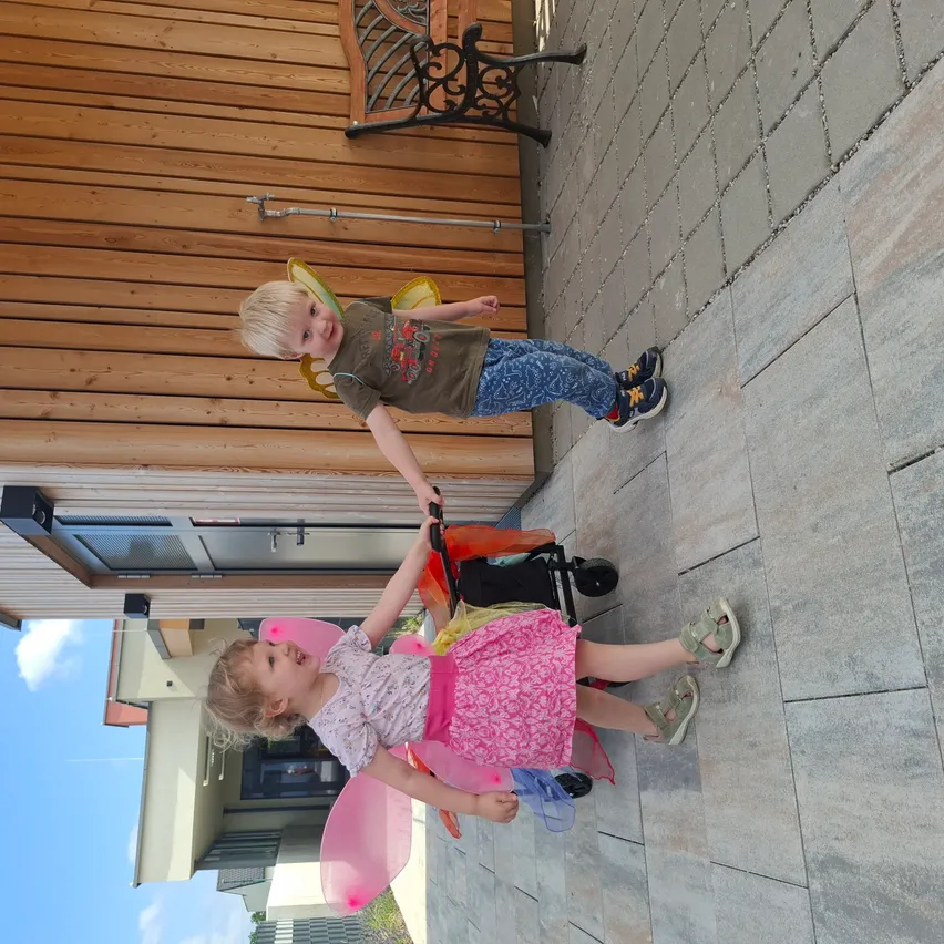 Zwei Kinder, ein Junge und ein Mädchen, stehen draußen. Der Junge ist in Flügeln eines Feens gekleidet und hält einen Kinderwagen. Das Mädchen trägt ein rosa Feenkostüm und hält den Griff des Kinderwagens.