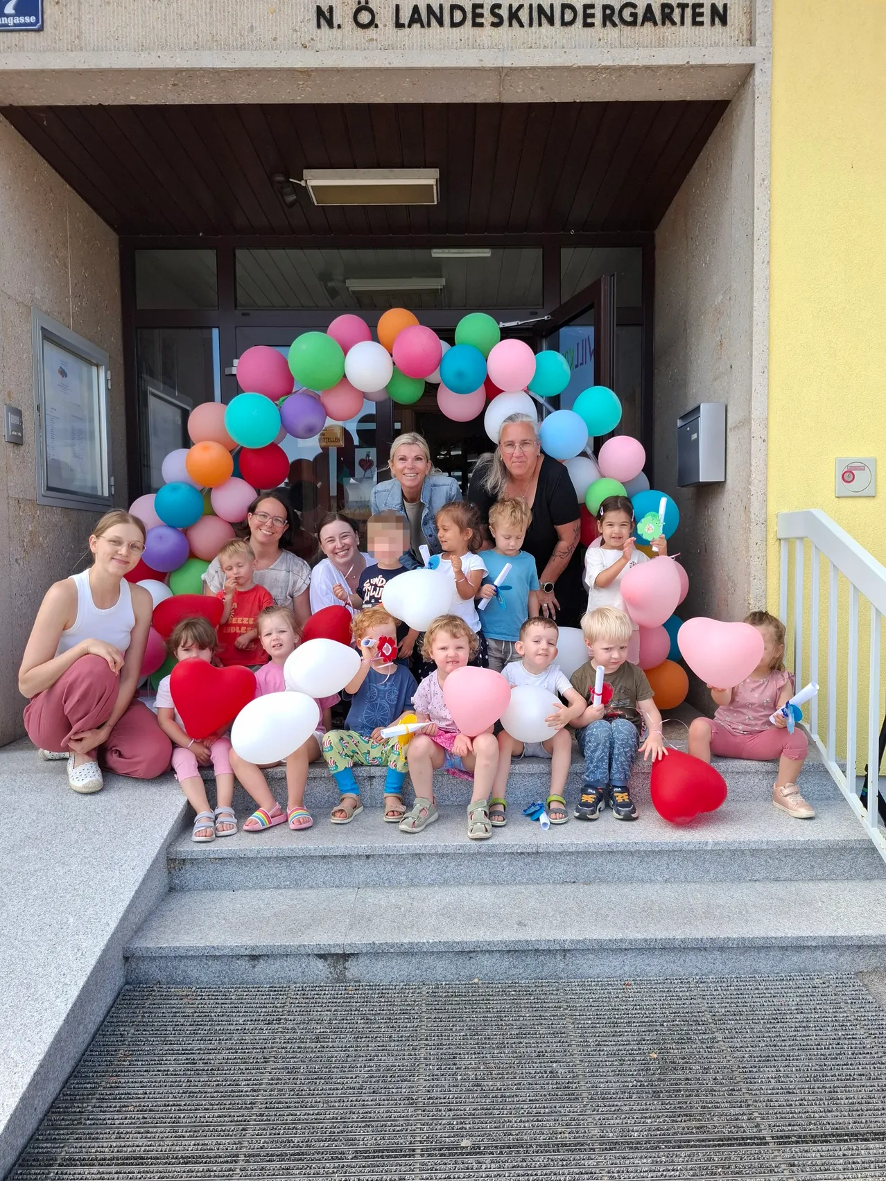 Eine Gruppe von Kindern und Erwachsenen posiert für ein Foto vor einem Gebäude und hält Luftballons und herzförmige Dekorationen. Die Umgebung umfasst einen farbenfrohen Ballonbogen und eine Treppe.