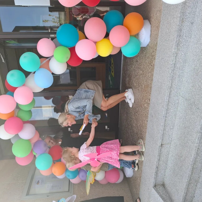 Eine Frau hält die Hand eines jungen Mädchens, beide stehen unter einem farbenfrohen Luftballonbogen vor einem Gebäude. Das Mädchen trägt ein Feenkostüm und die Frau hält einen Stift.