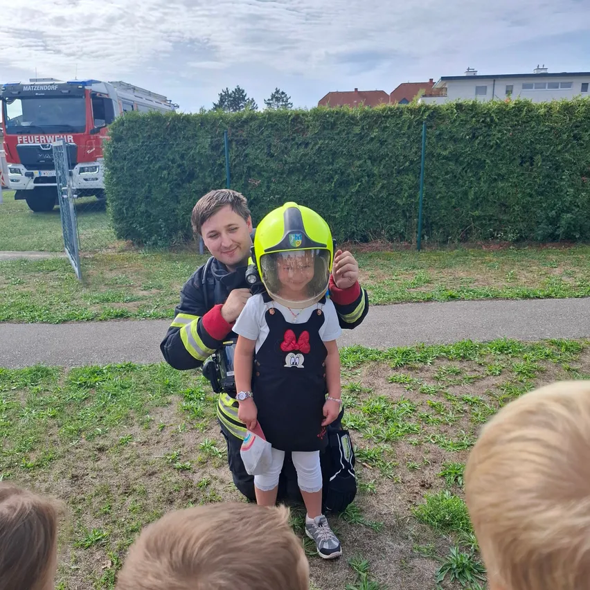 Ein Feuerwehrmann kniet und hält ein Kind, das einen Helm und ein Micky-Maus-Shirt trägt. Sie sind von Kindern umgeben und ein Feuerwehrwagen ist im Hintergrund zu sehen.